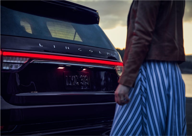 A person is shown near the rear of a 2023 Lincoln Aviator® SUV as the Lincoln Embrace illuminates the rear lights | Lincoln Demo 2 in Wooster OH