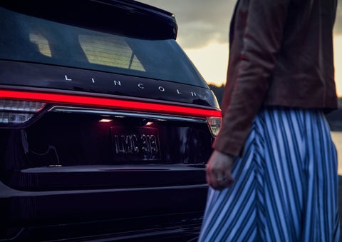 A person is shown near the rear of a 2024 Lincoln Aviator® SUV as the Lincoln Embrace illuminates the rear lights | Lincoln Demo 2 in Wooster OH